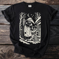Black t-shirt with a cream folk-cut print: a beaked Perchta in an apron sweeps a snowy doorstep, scissors in the basket on her back.