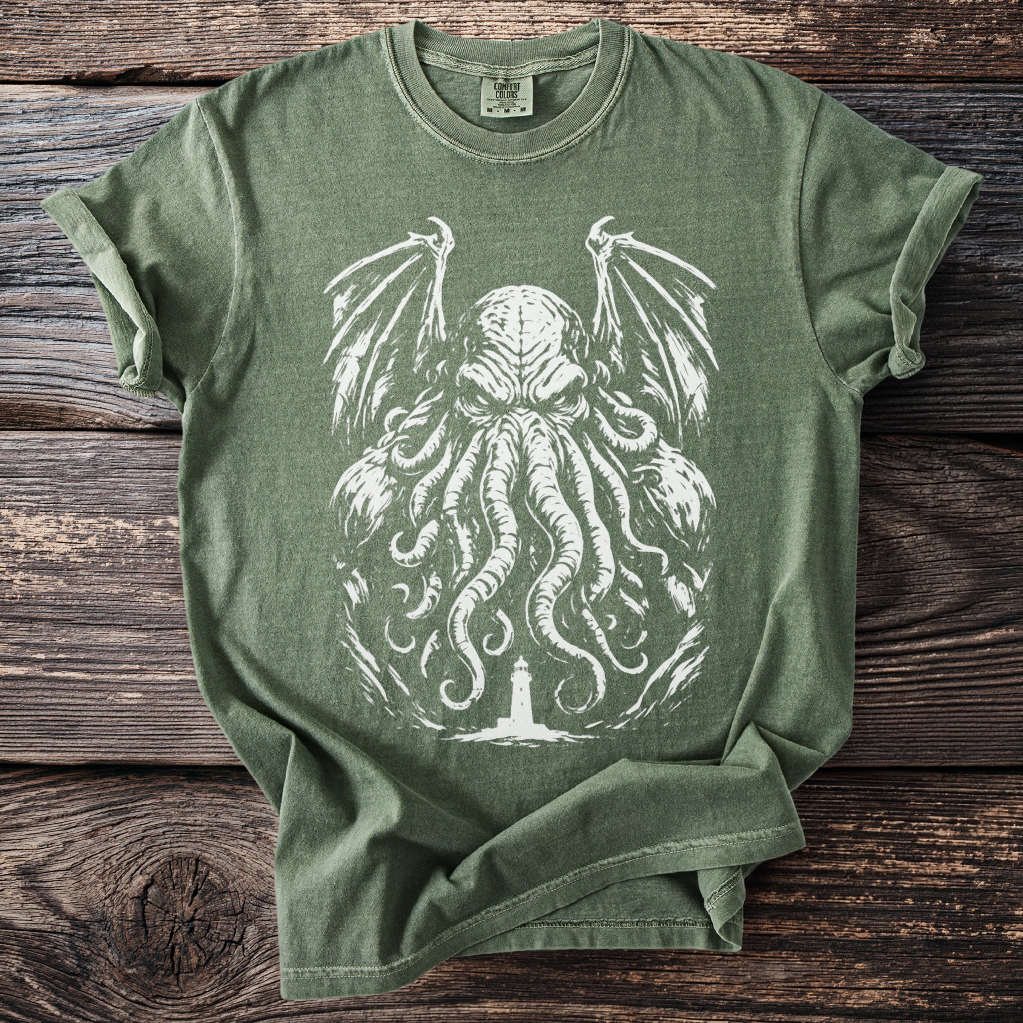 Green t-shirt with white tentacled creature and lighthouse graphic on wooden background.