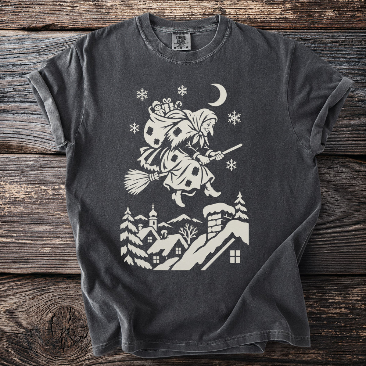 Pepper gray t-shirt with a cream folk-cut print: La Befana flies over snowy rooftops on her broom, a sack of gifts on her back.