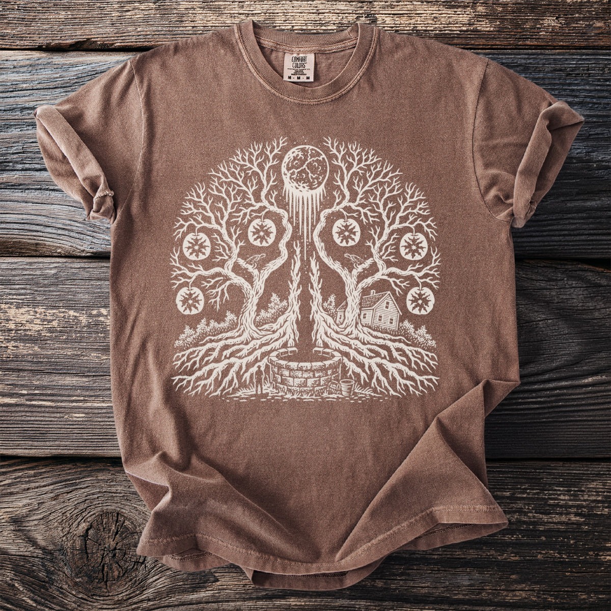 Brown t-shirt with white graphic of a tree with roots, moon, well, and house.