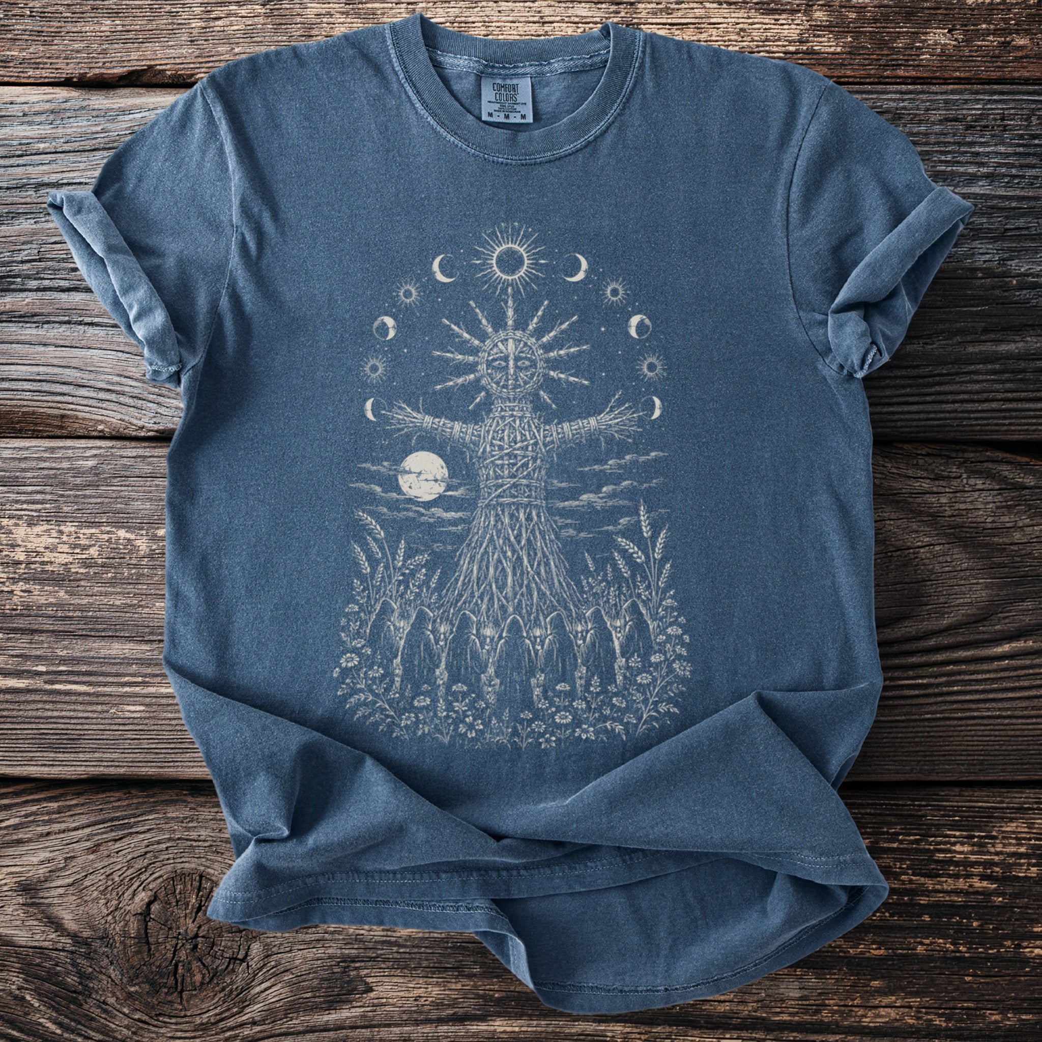 Blue t-shirt with a woven figure, sun, moons, and figures in a field.