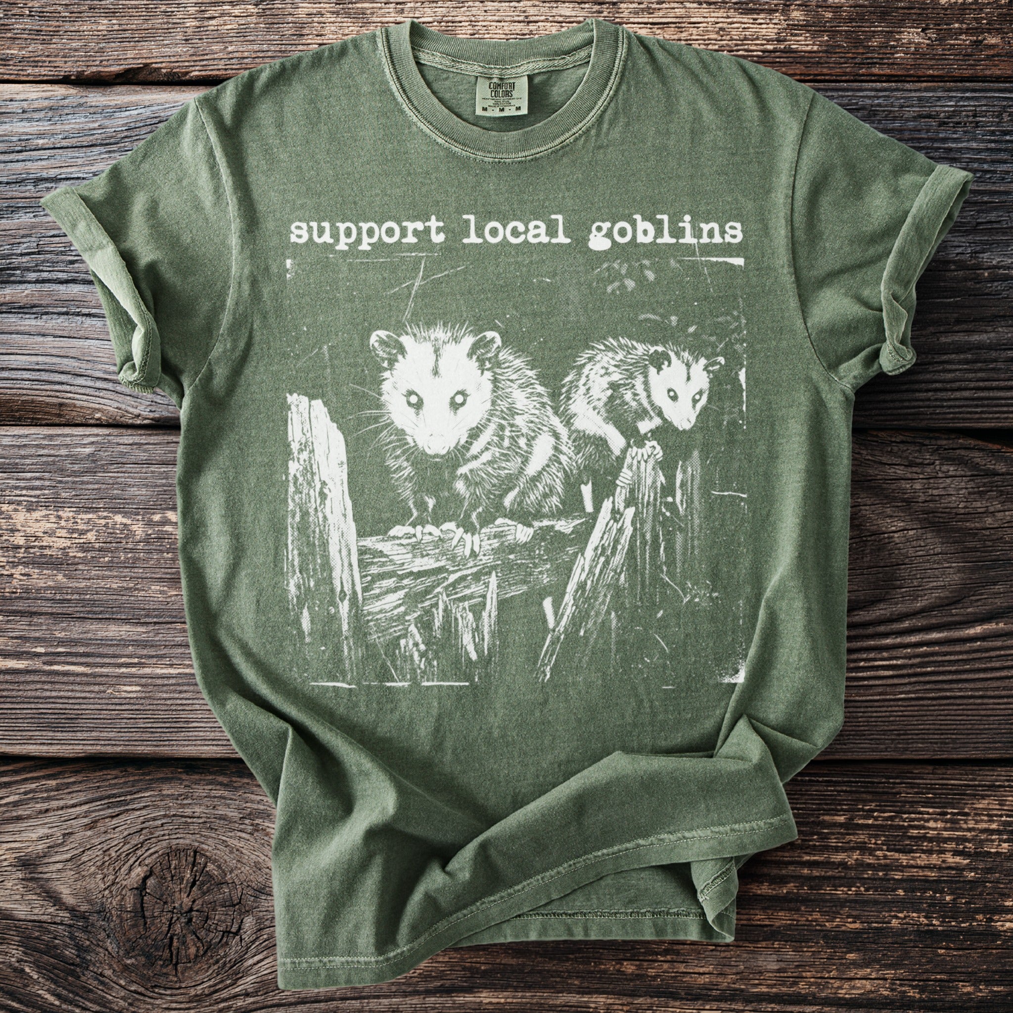 Comfort Colors green t-shirt with "support local goblins" and opossums on a fence.