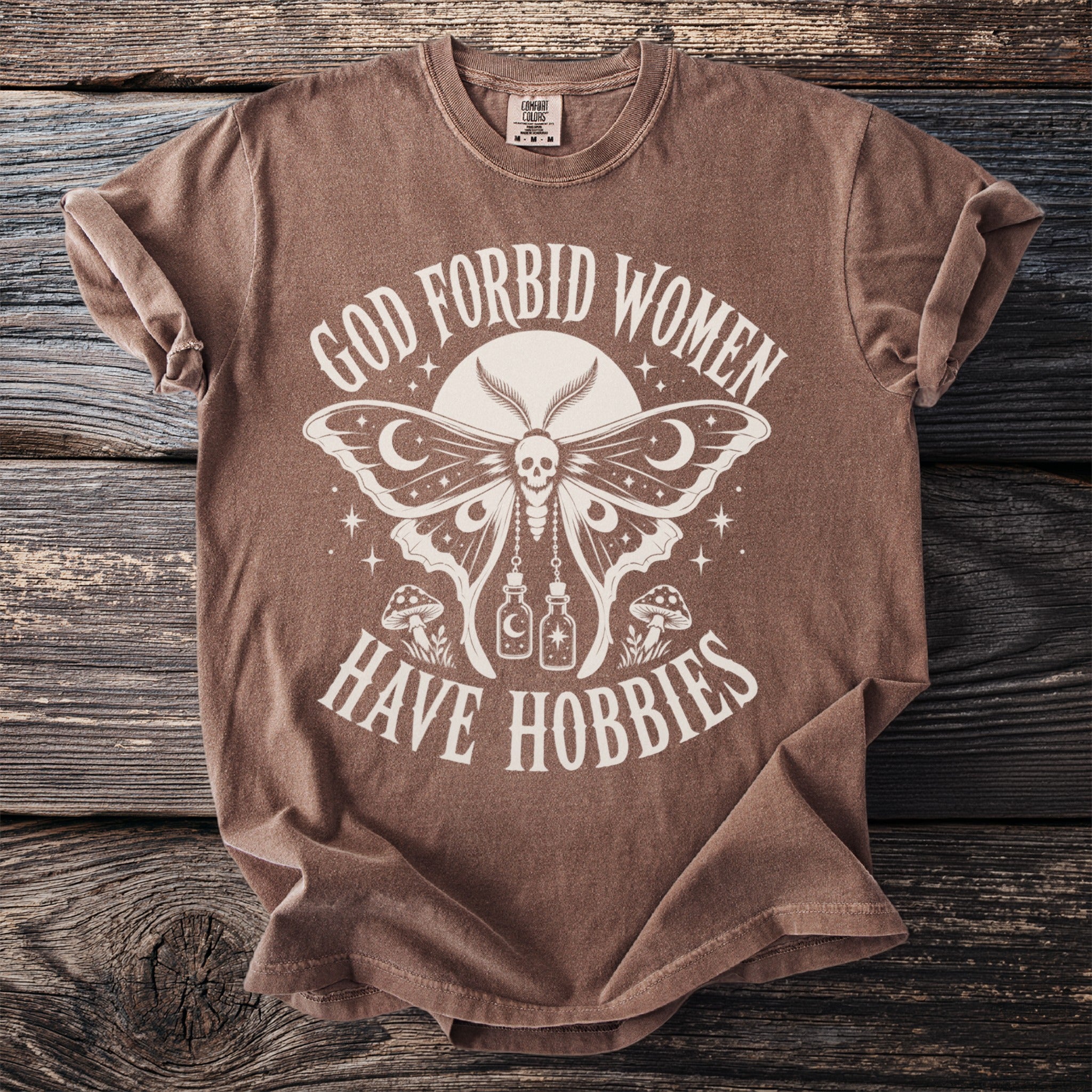 Brown t-shirt with text "God Forbid Women Have Hobbies" and moth graphic.