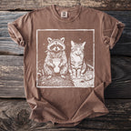 Brown t-shirt with print of raccoon holding drink and cat smoking, on wooden background.