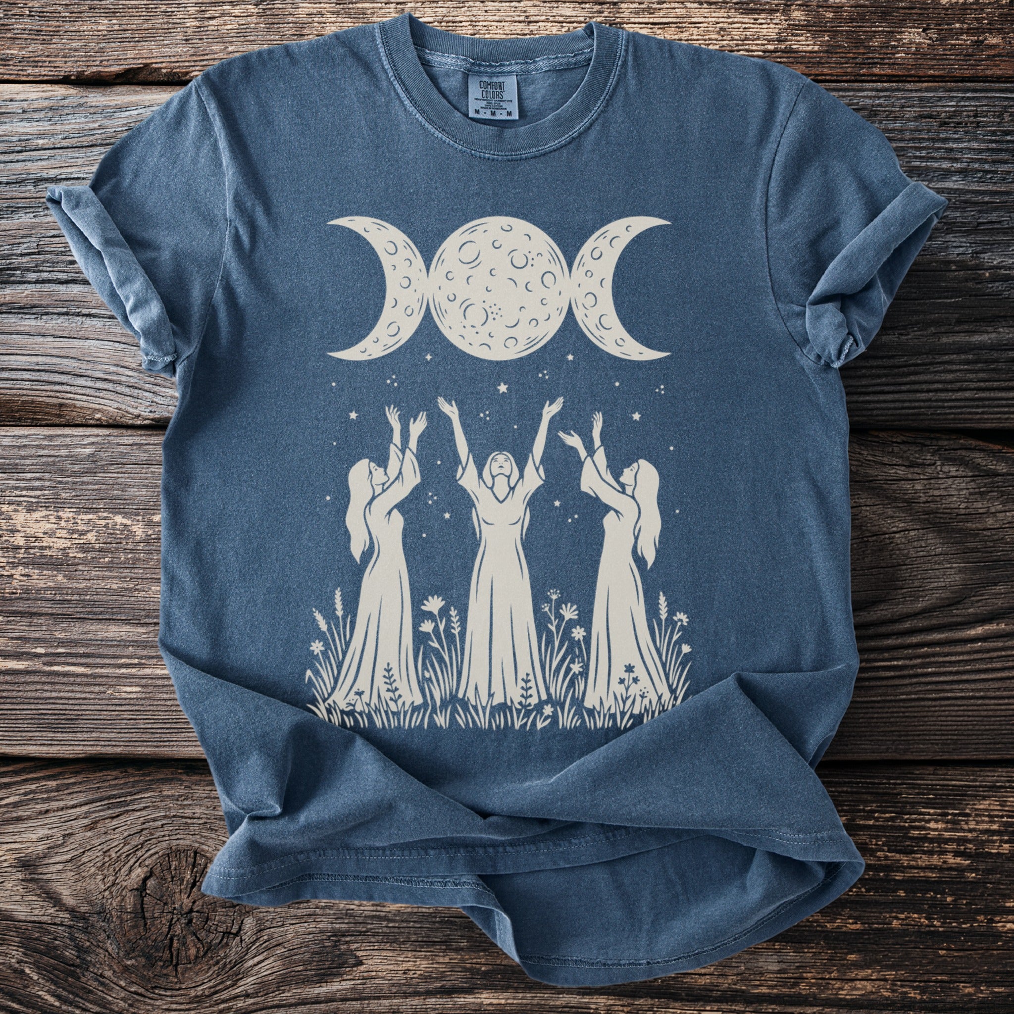 Blue t-shirt with a cream graphic of women reaching for a triple moon.