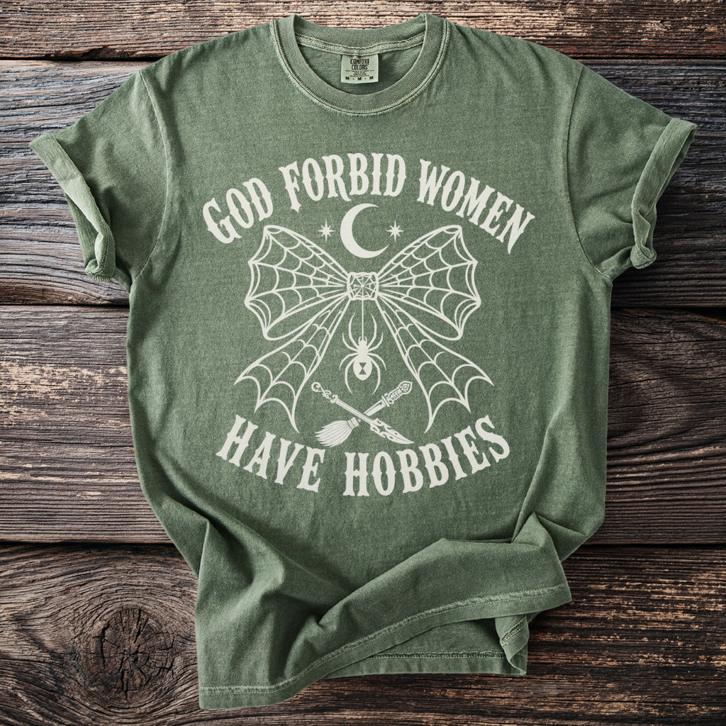 Green t-shirt featuring "God Forbid Women Have Hobbies" with spiderweb, spider, broom, and knife graphic.