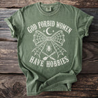Green t-shirt featuring "God Forbid Women Have Hobbies" with spiderweb, spider, broom, and knife graphic.