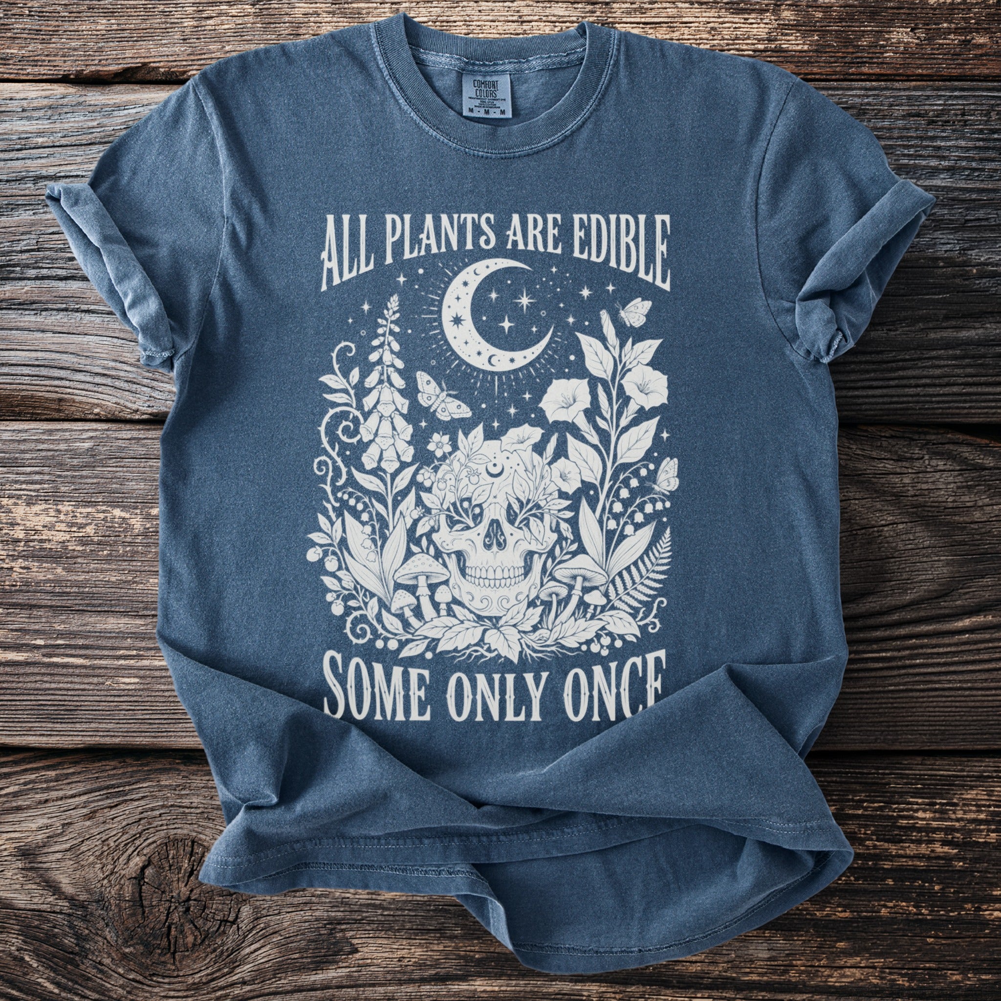Blue t-shirt with a skull, crescent moon, plants, and text "ALL PLANTS ARE EDIBLE SOME ONLY ONCE".