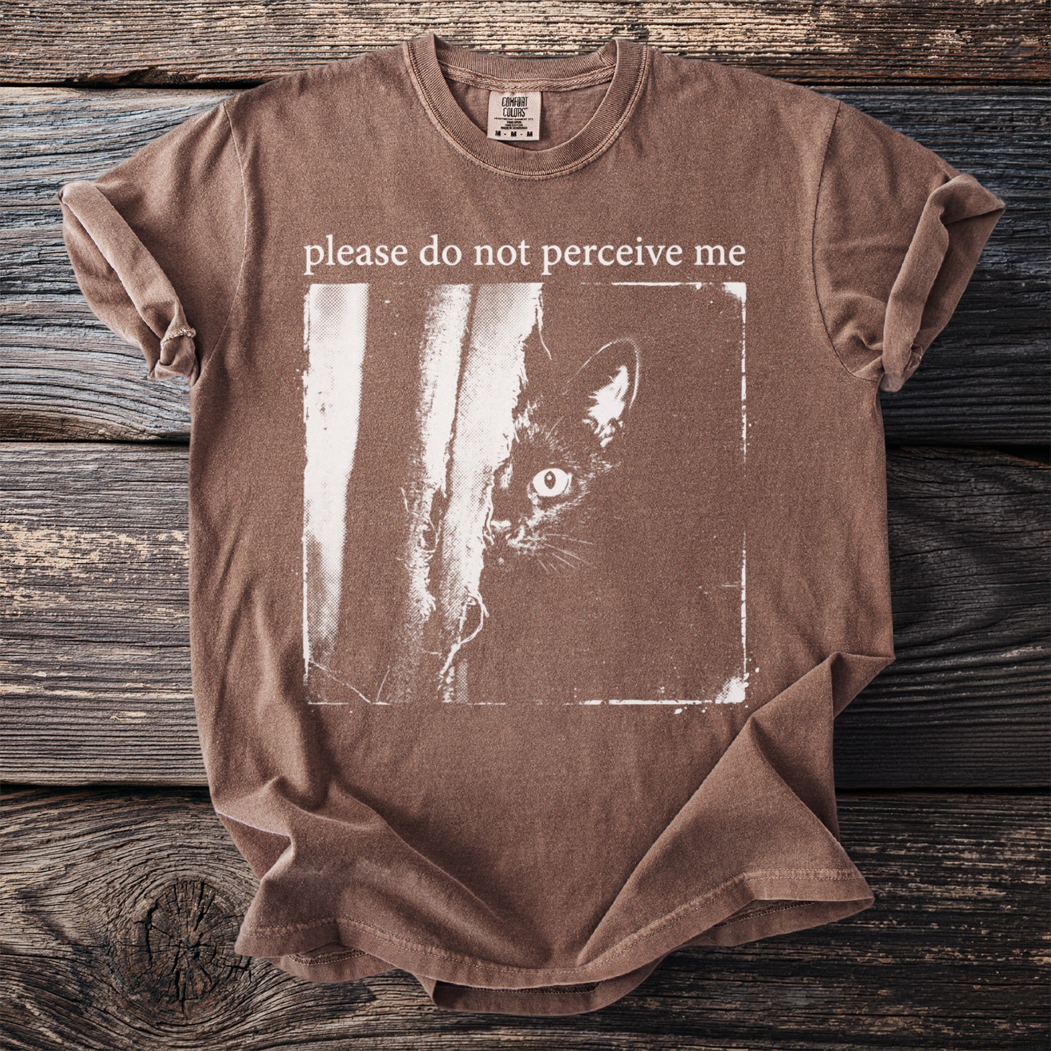 Brown t-shirt with white text "please do not perceive me" and a black cat peeking.