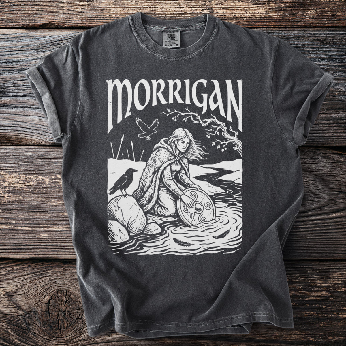 Dark gray t-shirt with "MORRIGAN" text and illustration of a woman by a river with a raven.