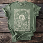 Green t-shirt with a cream-colored print of a queen spinning yarn with birds and constellations.