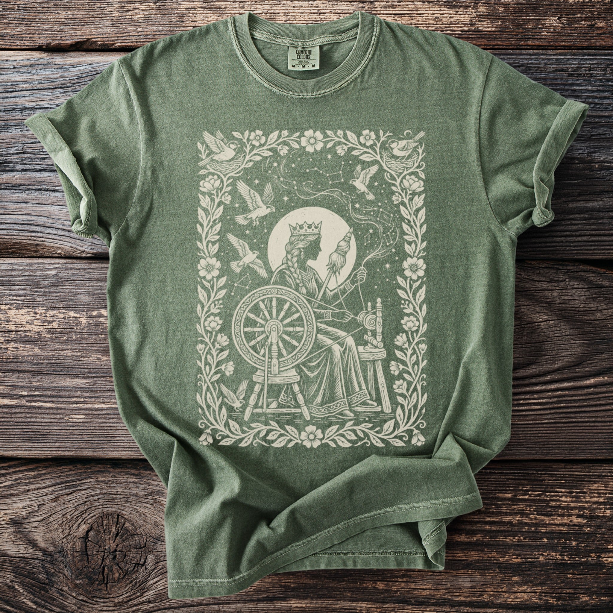Green t-shirt with a cream-colored print of a queen spinning yarn with birds and constellations.