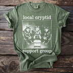 Green t-shirt with "local cryptid support group" text and image of three wolves.