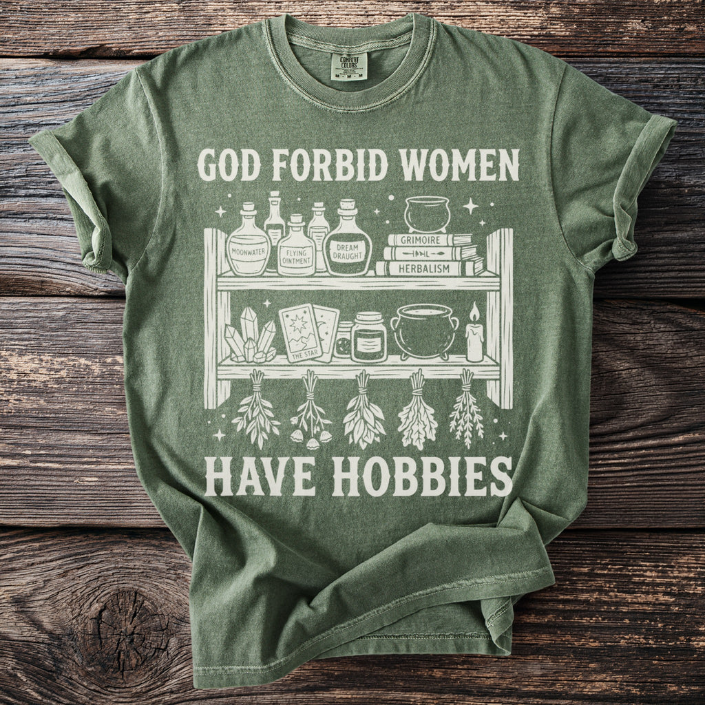 Green t-shirt with white graphic of shelves holding potion bottles, books, crystals, tarot cards, and herbs. Text says "GOD FORBID WOMEN HAVE HOBBIES".