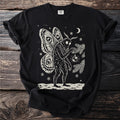 Black t-shirt with a cream folk-cut print: Mothman, eyespot wings open, hangs one glass ornament on a pine branch.