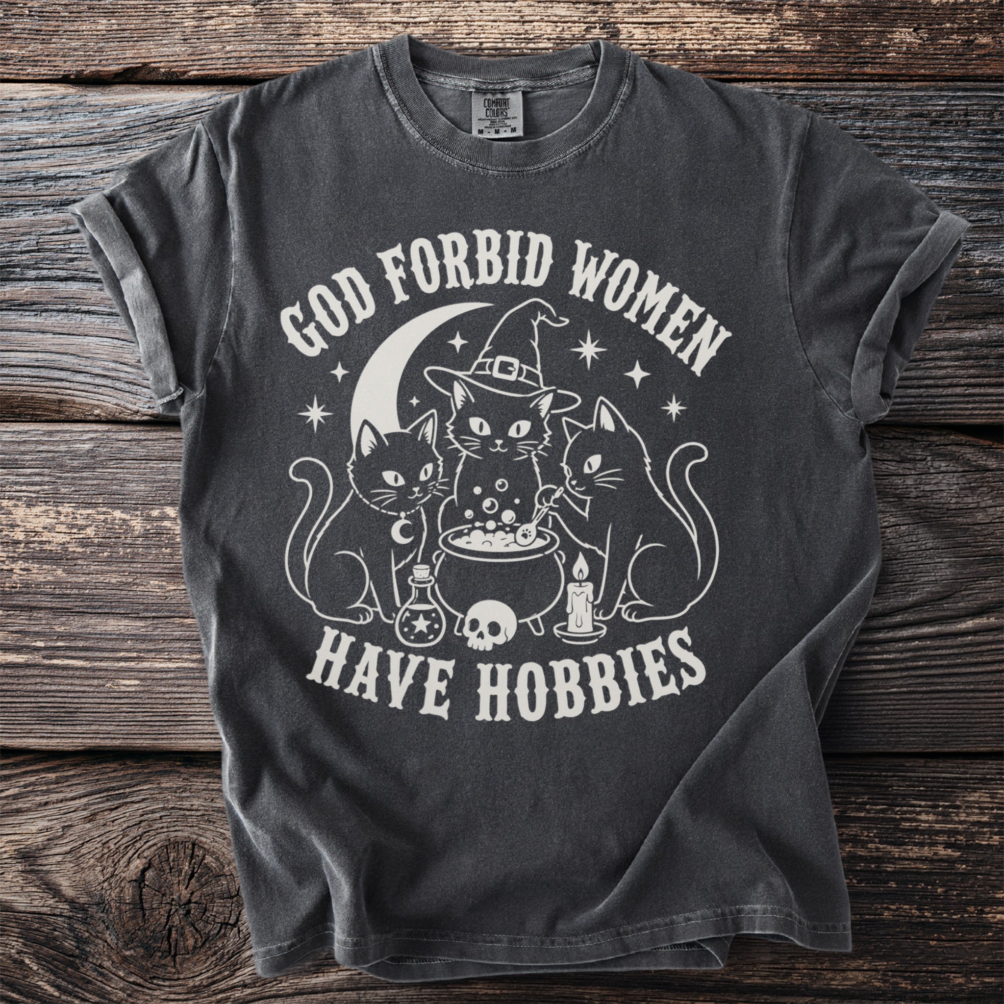 Dark gray t-shirt with "God Forbid Women Have Hobbies" text and witchy cat graphic.