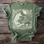 Green t-shirt with a cream colored linocut design of a fox and raven surrounded by foliage and mushrooms.