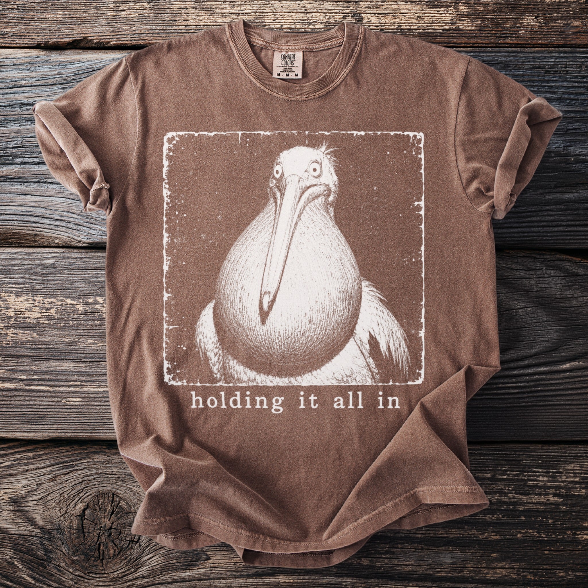 Brown t-shirt with a distressed graphic of a pelican and the text "holding it all in".