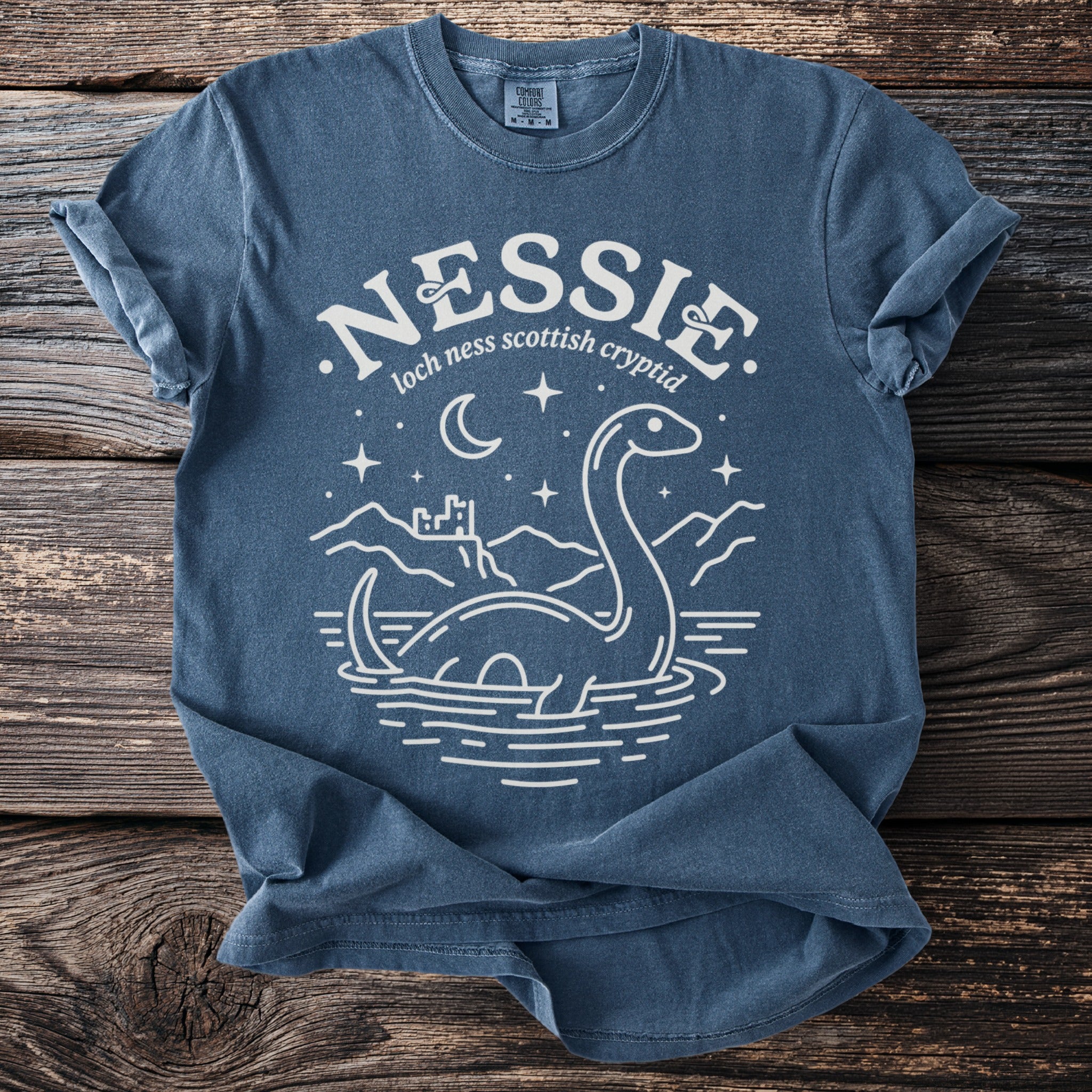 Blue t-shirt with white graphic of Nessie, castle, moon, and stars.