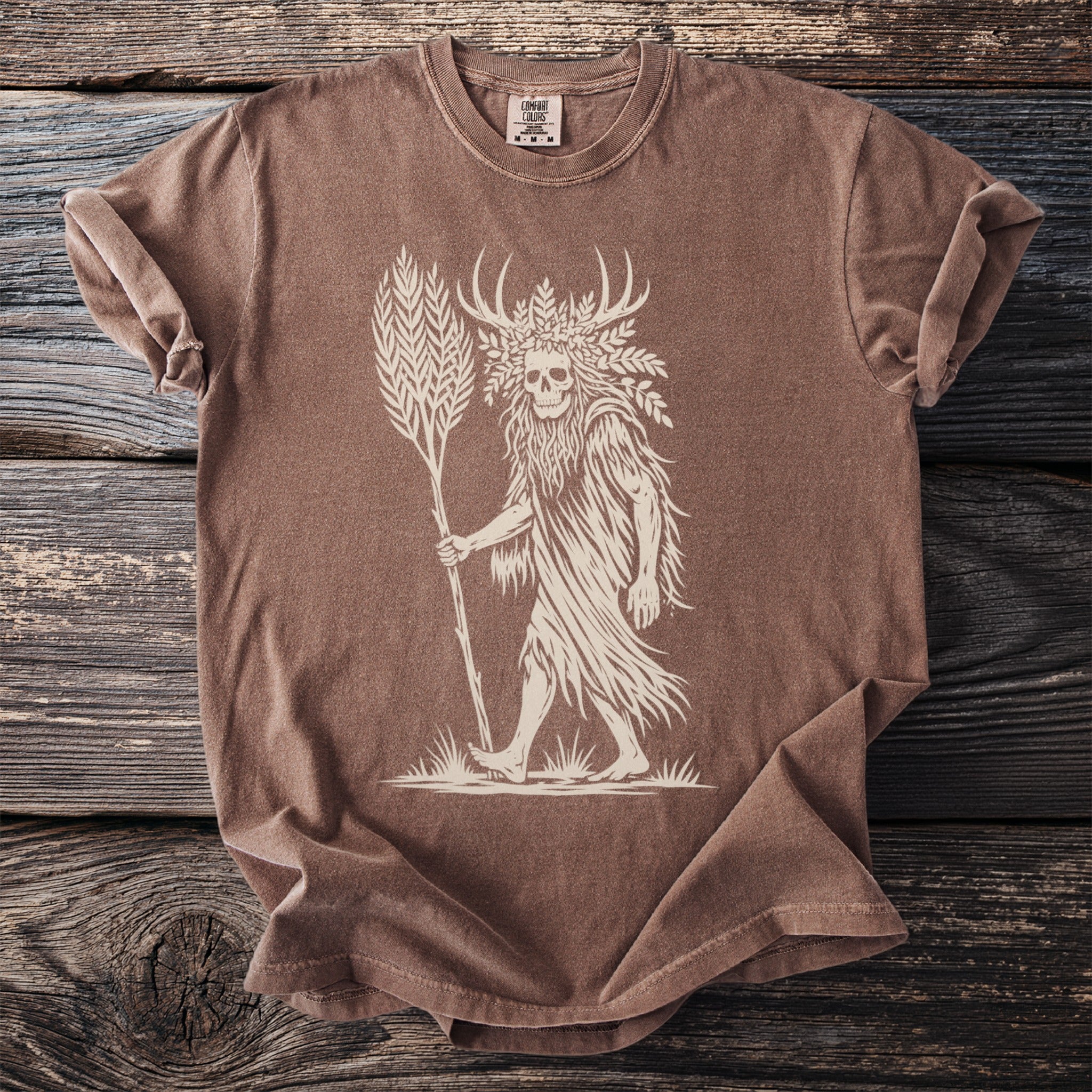 Brown t-shirt with a cream graphic of a skull-headed figure with antlers holding a stalk of wheat.