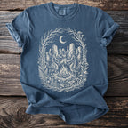 Blue t-shirt with a graphic of hooded figures dancing around a bonfire under a moonlit sky.