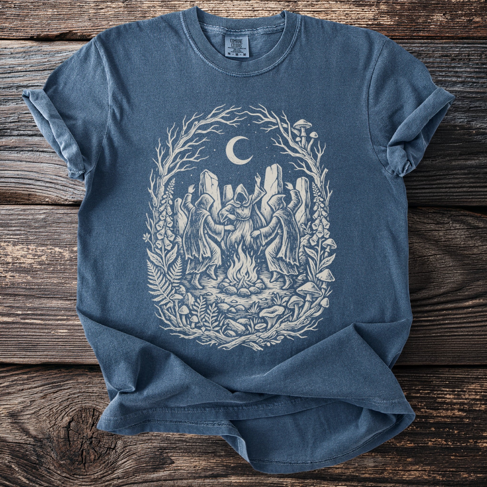 Blue t-shirt with a graphic of hooded figures dancing around a bonfire under a moonlit sky.
