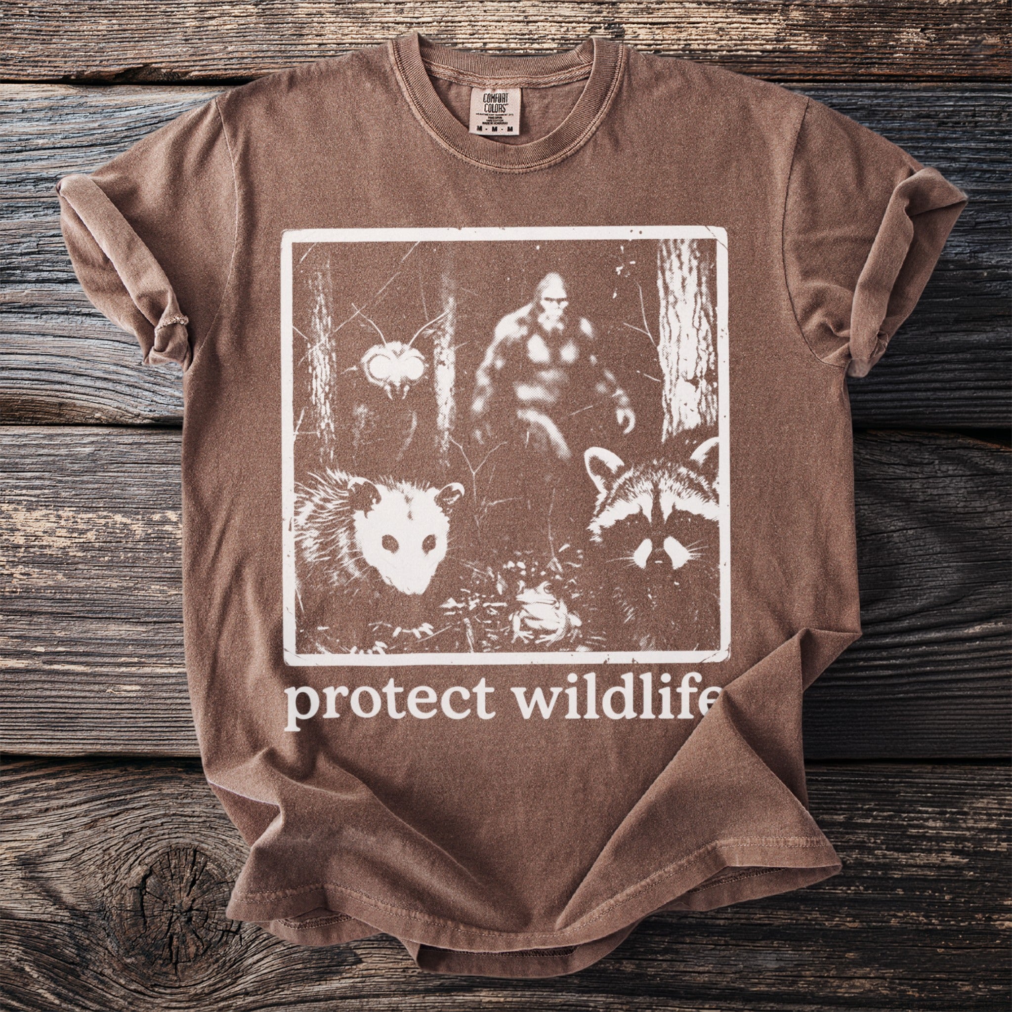 Brown t-shirt with graphic of Bigfoot, opossum, raccoon, insect, and frog. Text reads "protect wildlife".
