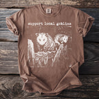 Brown t-shirt with white print of two possums on a wooden fence and text "support local goblins".