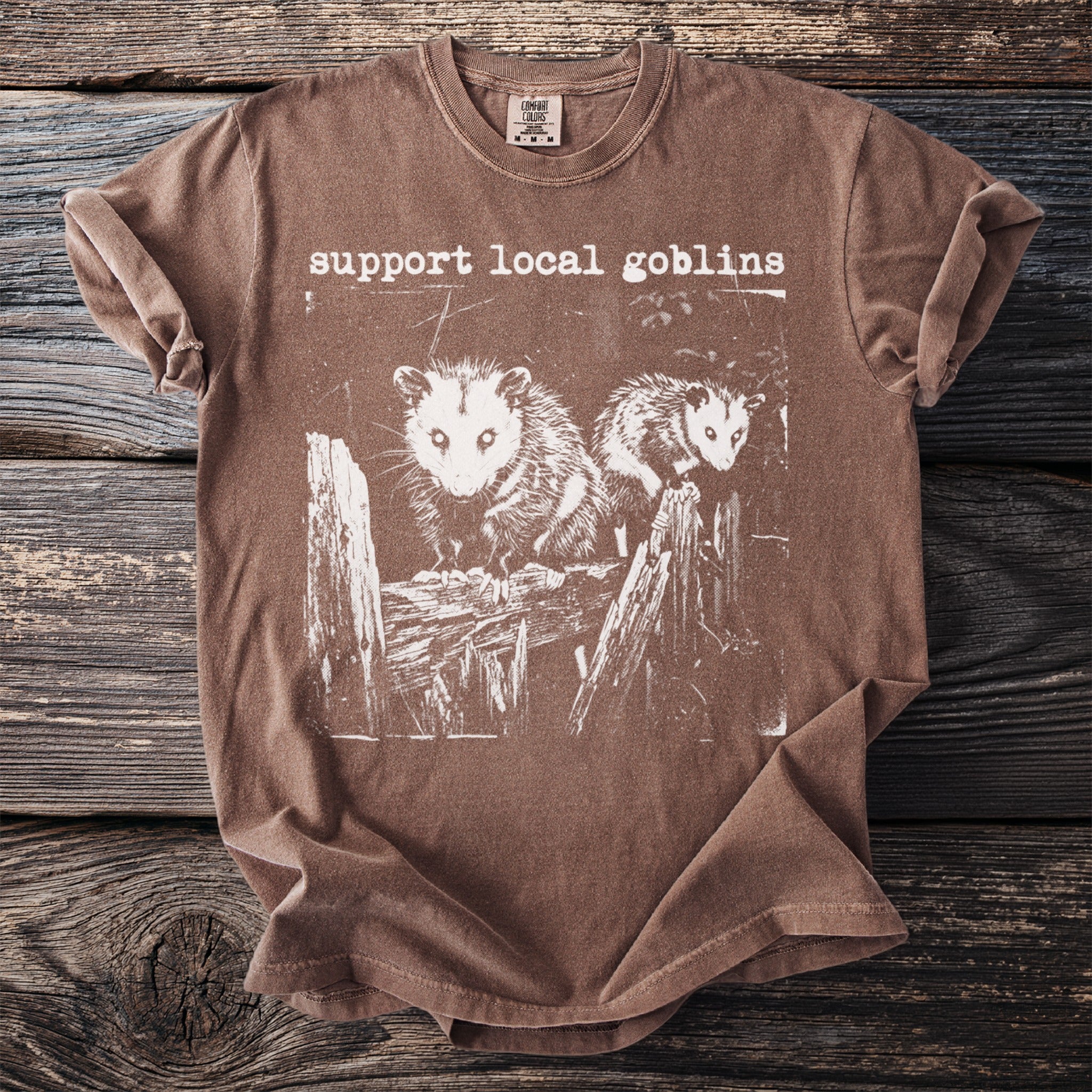 Brown t-shirt with white print of two possums on a wooden fence and text "support local goblins".