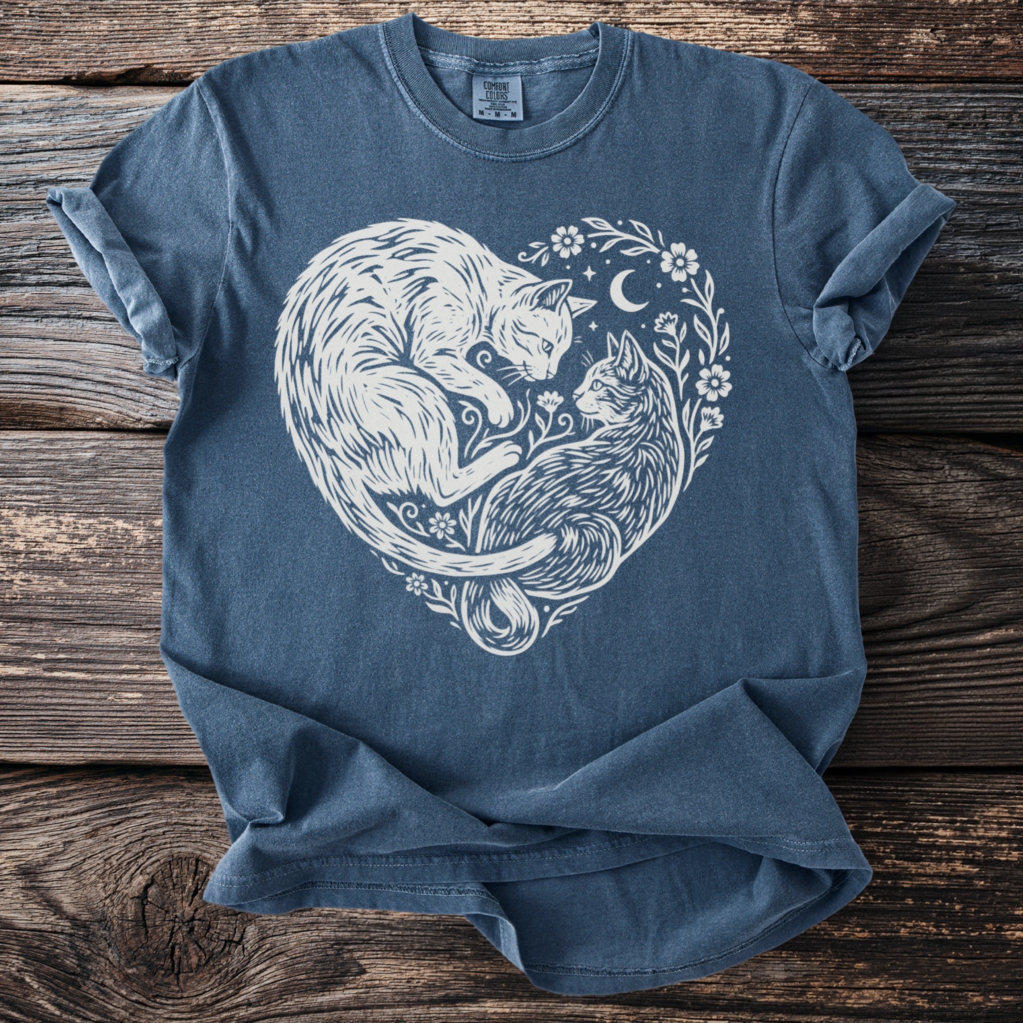 Blue t-shirt with white linework art of two cats forming a heart with floral accents and a moon.