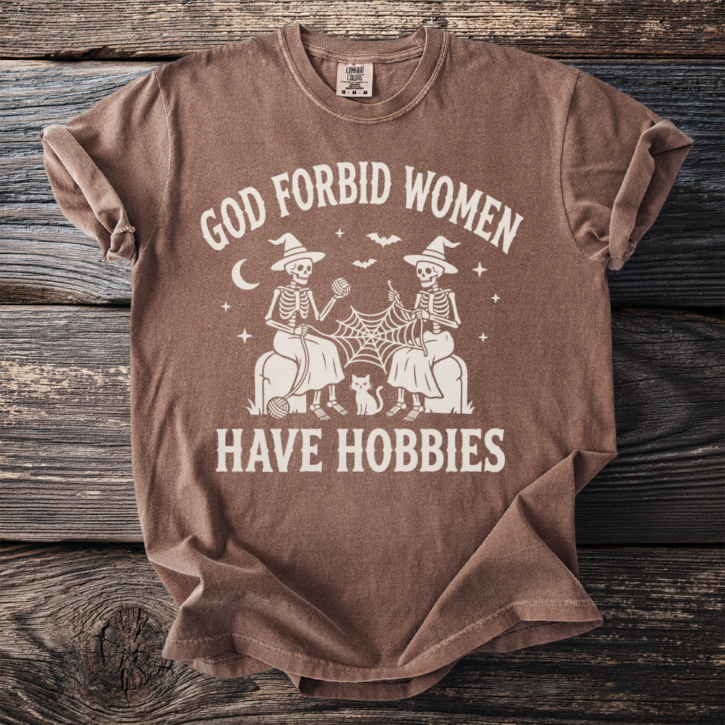 Brown t-shirt with skeletons knitting, cat, and text "GOD FORBID WOMEN HAVE HOBBIES."