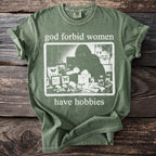 Green t-shirt with "god forbid women have hobbies" and a graphic of a person collecting insects.