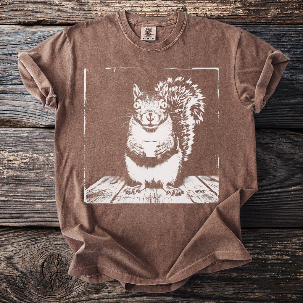 Brown t-shirt with a white graphic of a squirrel holding a cup on a wooden surface.