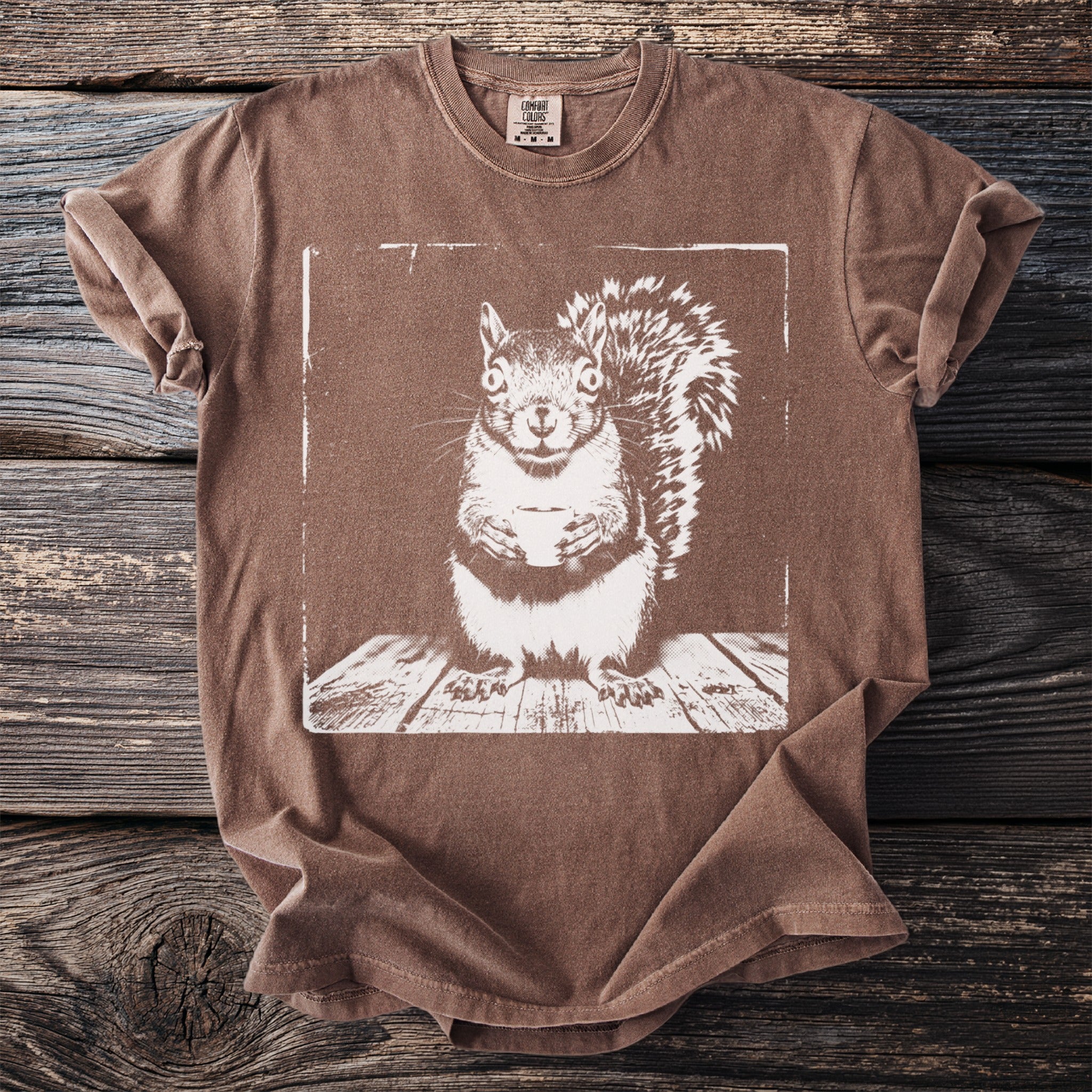 Brown t-shirt with a white graphic of a squirrel holding a cup on a wooden surface.
