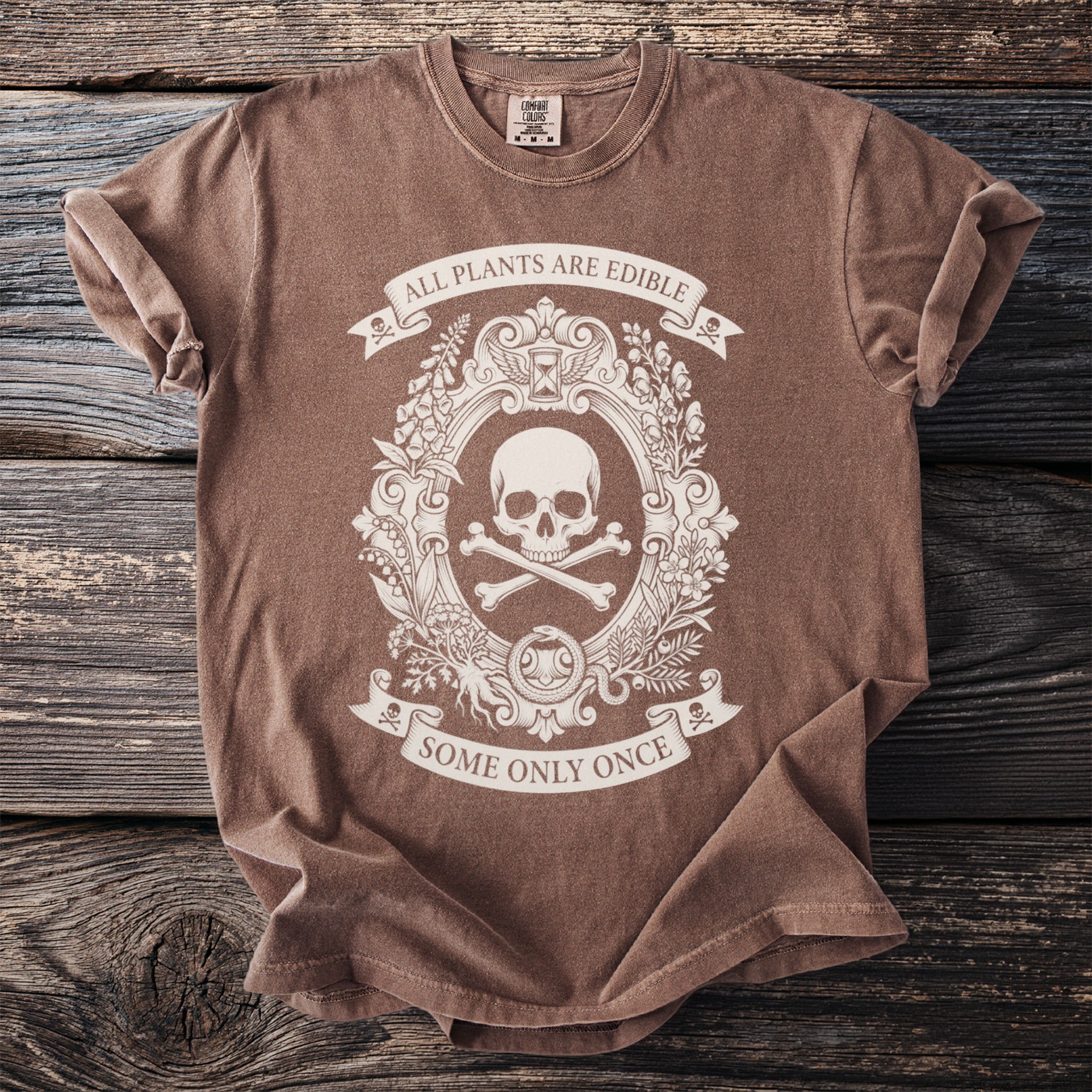 Brown t-shirt with a cream oval graphic of a skull and crossbones, botanical elements, and text.