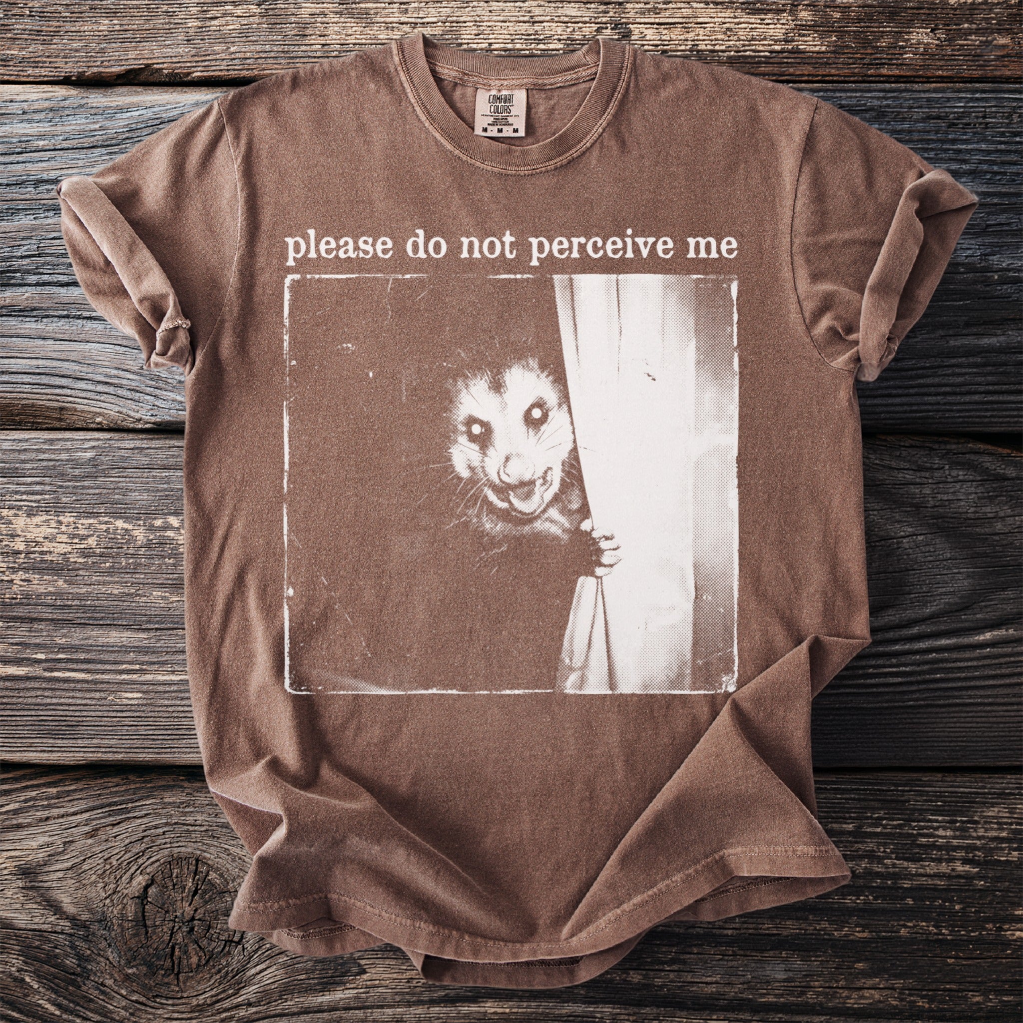 Brown t-shirt with a graphic of an opossum behind a curtain and text "please do not perceive me".