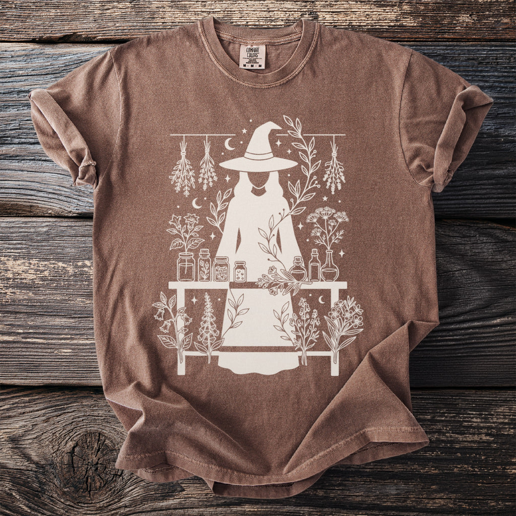 Brown t-shirt with a white line drawing of a witch and botanical elements.