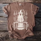 Brown t-shirt with a white line drawing of a witch and botanical elements.