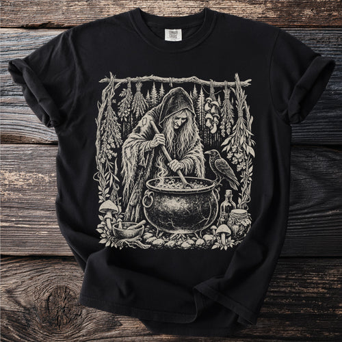 Black t-shirt featuring an illustration of a witch stirring a cauldron with a raven nearby.