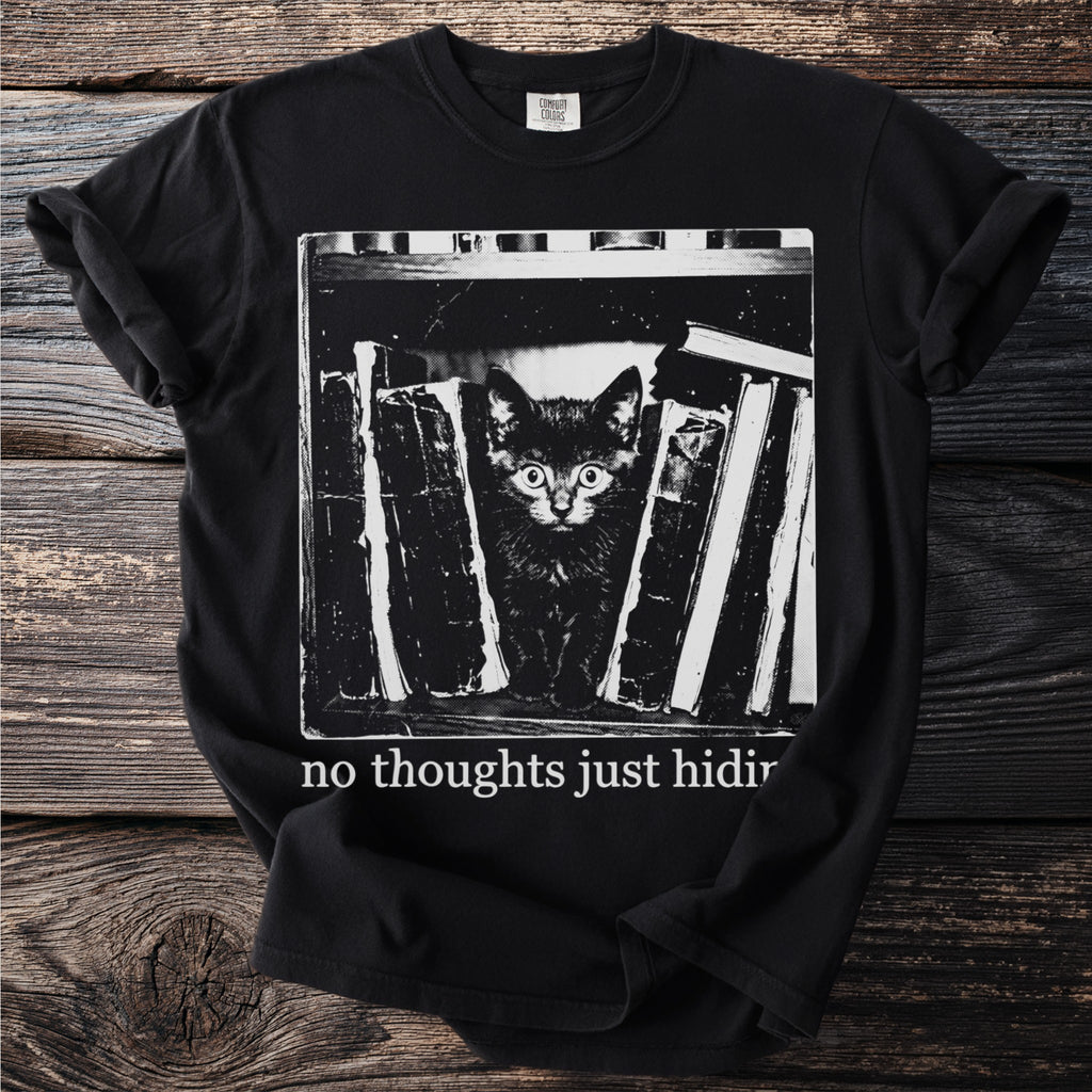 Black t-shirt with a kitten peeking from behind books with text "no thoughts just hiding".