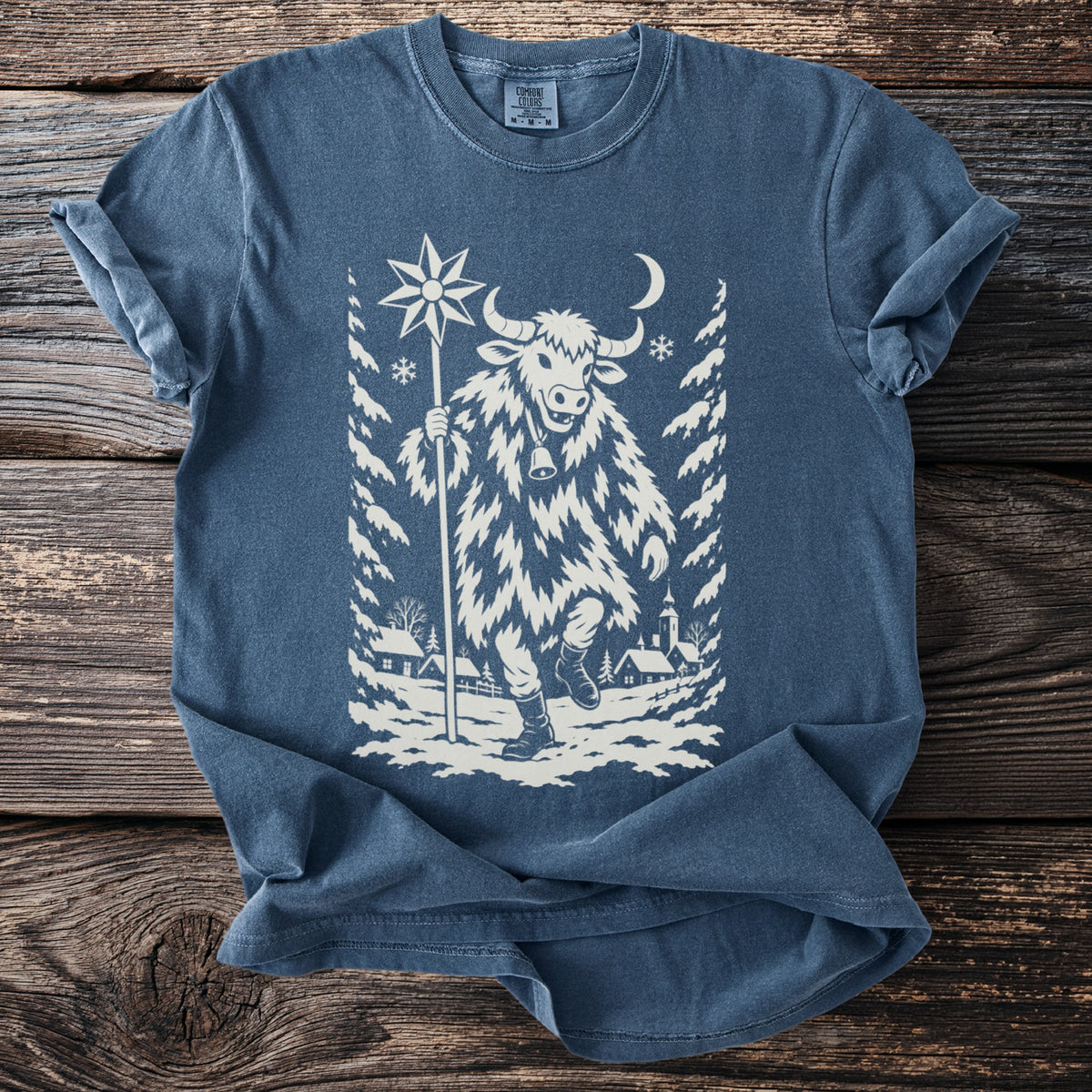 Denim blue t-shirt with a cream folk-cut print: a bull-masked figure in a shaggy coat walks through a snowy village carrying a star on a pole.