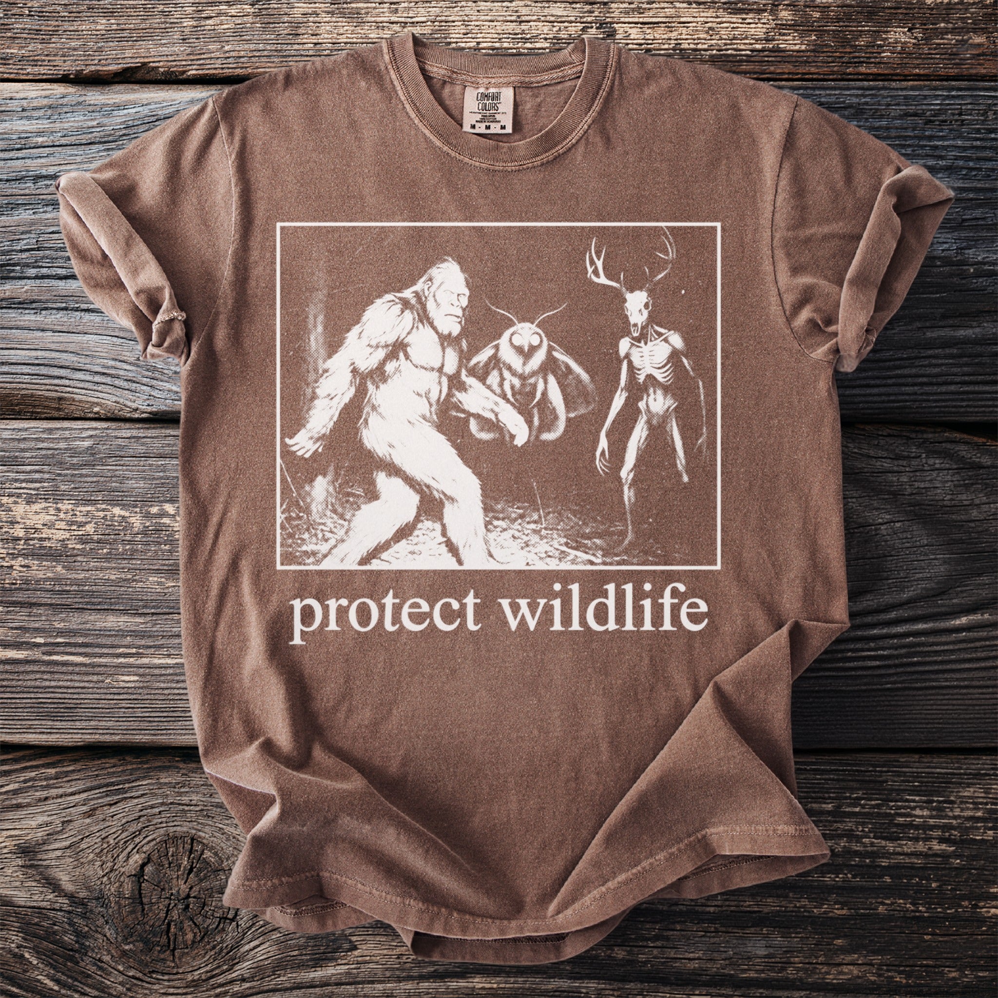 Brown t-shirt with Bigfoot, moth, and deer skull creatures and "protect wildlife" text.
