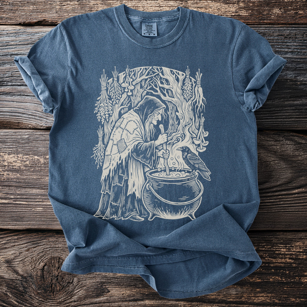 Blue t-shirt with a graphic of a witch stirring a cauldron with a raven nearby.