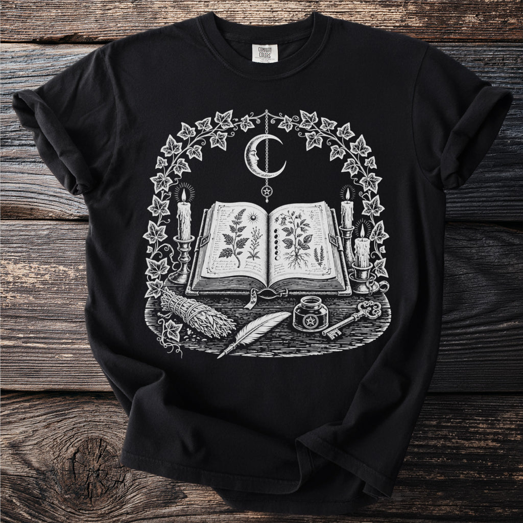 Black t-shirt with an open book, moon, candles, herbs, feather, ink, and key.