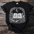 Black t-shirt with an open book, moon, candles, herbs, feather, ink, and key.