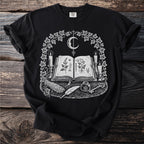 Black t-shirt with an open book, moon, candles, herbs, feather, ink, and key.