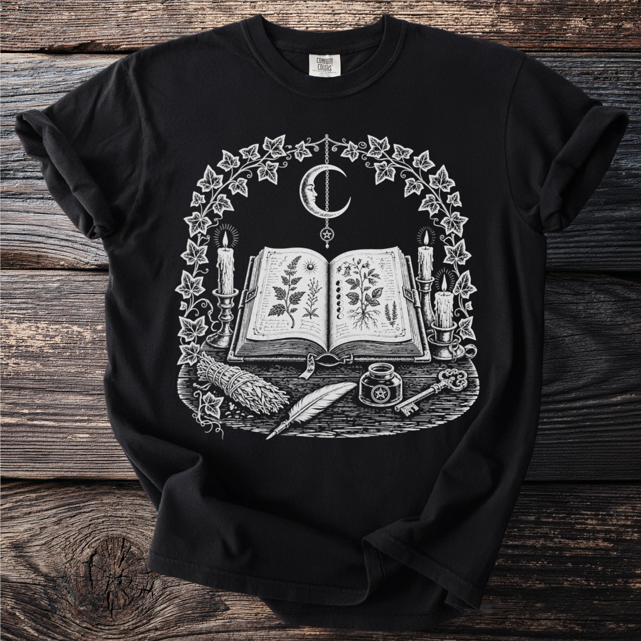 Black t-shirt with an open book, moon, candles, herbs, feather, ink, and key.