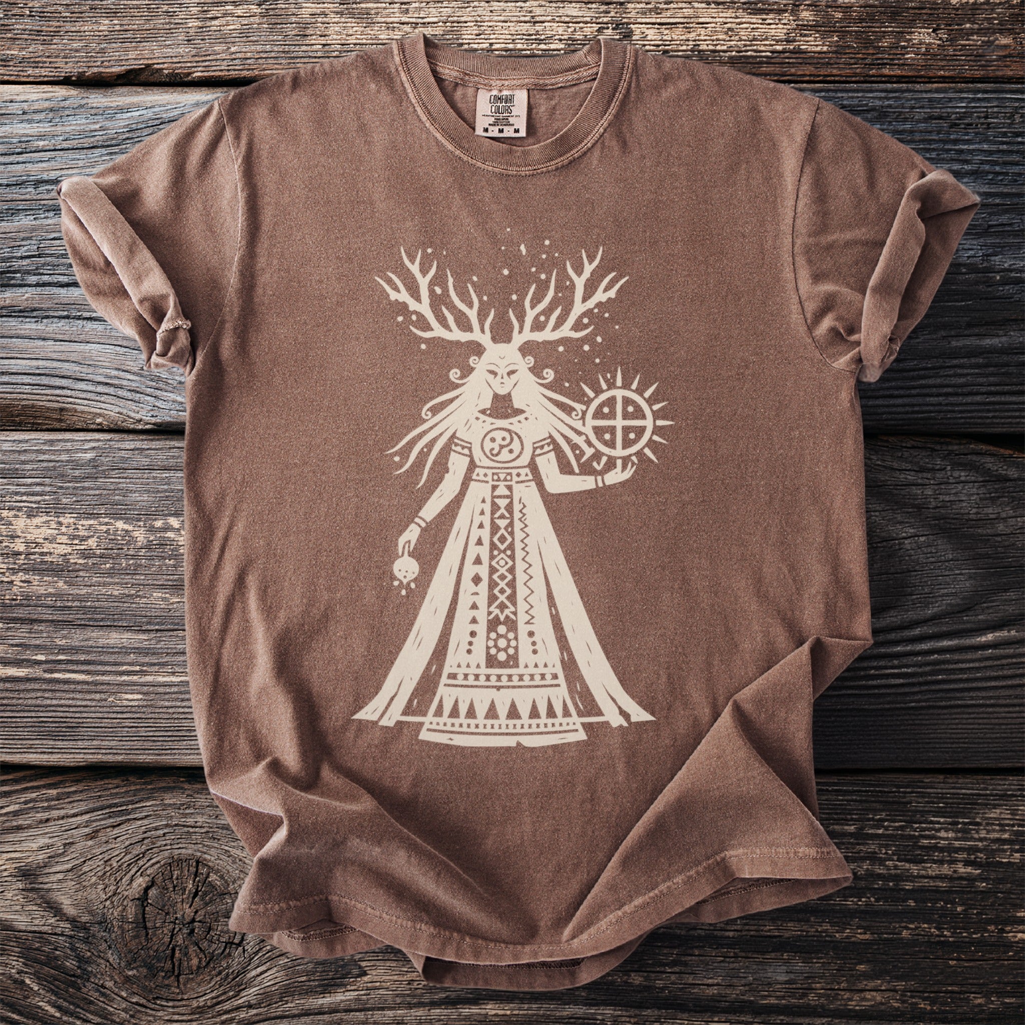 Brown Comfort Colors t-shirt with a cream graphic of a horned figure holding a sun-like symbol.