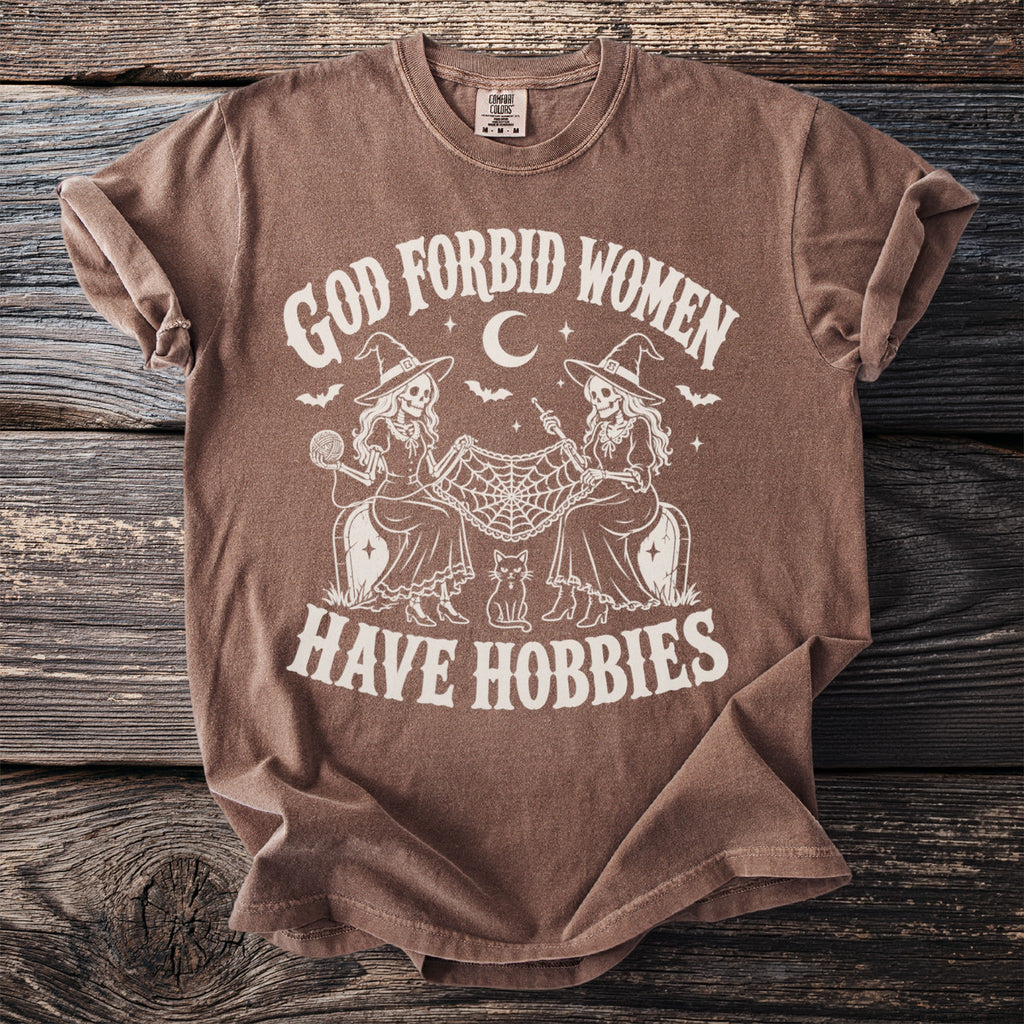 Brown t-shirt with "God Forbid Women Have Hobbies" text featuring two skeleton witches.