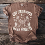 Brown t-shirt with "God Forbid Women Have Hobbies" text featuring two skeleton witches.